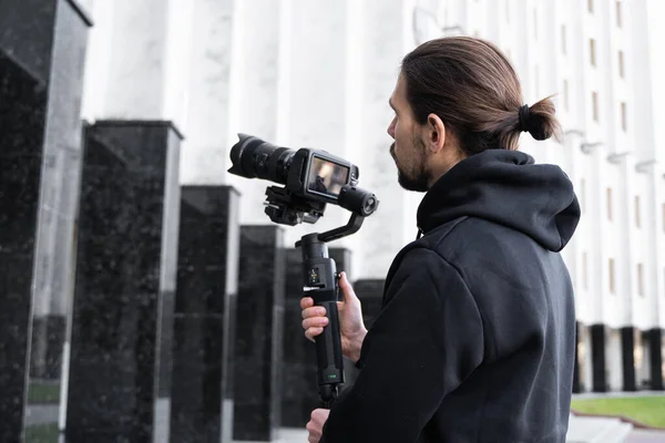 Young Professional videographer holding professional camera on 3-axis gimbal stabilizer. Pro equipment helps to make high quality video without shaking. Cameraman wearing black hoodie making a videos.