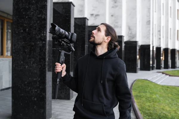 Young Professional videographer holding professional camera on 3-axis gimbal stabilizer. Pro equipment helps to make high quality video without shaking. Cameraman wearing black hoodie making a videos.