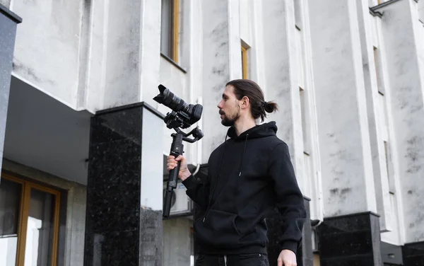Ung professionell videograf som håller professionell kamera på 3-axlig gimbal stabilisator. Pro-utrustning hjälper till att göra högkvalitativ video utan att skaka. Kameraman bär svart luvtröja gör en video. — Stockfoto