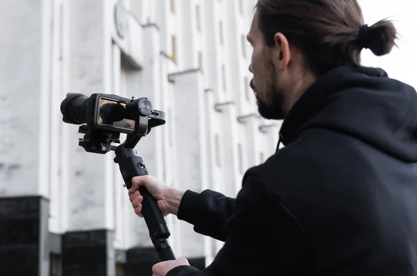 Ung professionell videograf som håller professionell kamera på 3-axlig gimbal stabilisator. Pro-utrustning hjälper till att göra högkvalitativ video utan att skaka. Kameraman bär svart luvtröja gör en video. — Stockfoto