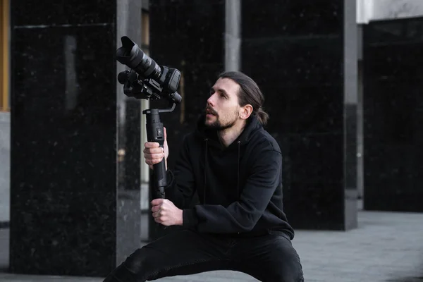 Young Professional videographer holding professional camera on 3-axis gimbal stabilizer. Pro equipment helps to make high quality video without shaking. Cameraman wearing black hoodie making a videos.