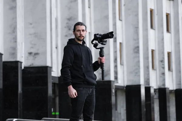 Młody profesjonalny fotograf trzymający profesjonalną kamerę na 3-osiowym stabilizatorze gimbalu. Pro sprzęt pomaga w tworzeniu wysokiej jakości wideo bez wstrząsania. Kamerzysta w czarnej bluzie kręci filmy. — Zdjęcie stockowe