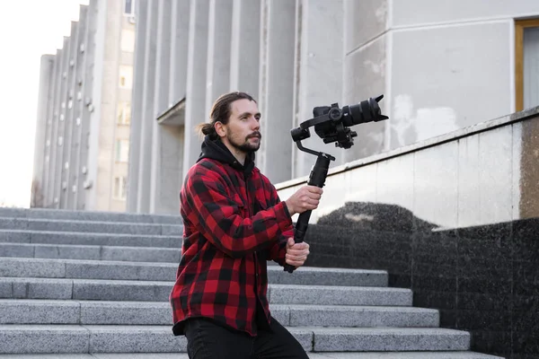 Giovane videografo professionale con fotocamera professionale su stabilizzatore gimbale a 3 assi. L'attrezzatura Pro aiuta a fare video di alta qualità senza scuotere. Cameraman indossa camicia rossa facendo un video . — Foto Stock