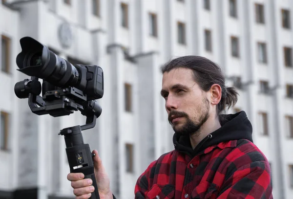 Jovem videógrafo profissional segurando câmera profissional no estabilizador de 3 eixos gimbal. Pro equipamento ajuda a fazer vídeo de alta qualidade sem tremer. Cameraman vestindo camisa vermelha fazendo um vídeos . — Fotografia de Stock