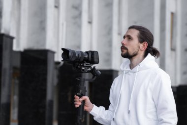 3 eksenli titreşimli dengeleyicide profesyonel kamera tutan genç profesyonel kameraman. Pro ekipmanlar titremeden yüksek kaliteli video çekmeye yardımcı olur. Beyaz kapüşonlu bir kameraman video çekiyor..