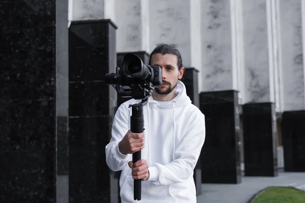 Jovem videógrafo profissional segurando câmera profissional no estabilizador de 3 eixos gimbal. Pro equipamento ajuda a fazer vídeo de alta qualidade sem tremer. Cameraman vestindo capuz branco fazendo um vídeo . — Fotografia de Stock