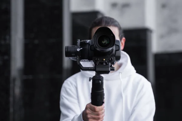 3軸ジンバルスタビライザーにプロのカメラを保持している若いプロのビデオカメラマン.プロ機器は、揺れずに高品質のビデオを作成するのに役立ちます。カメラマン身に着けていますホワイトパーカー作るビデオ. — ストック写真