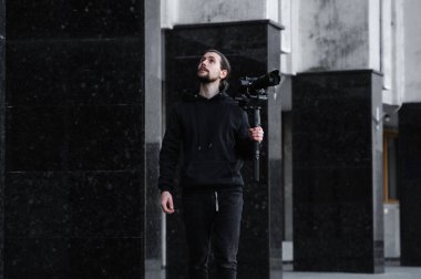3 eksenli titreşimli dengeleyicide profesyonel kamera tutan genç profesyonel kameraman. Pro ekipmanlar titremeden yüksek kaliteli video çekmeye yardımcı olur. Siyah kapüşonlu bir kameraman video çekiyor..