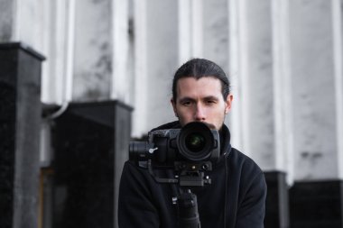 3 eksenli titreşimli dengeleyicide profesyonel kamera tutan genç profesyonel kameraman. Pro ekipmanlar titremeden yüksek kaliteli video çekmeye yardımcı olur. Siyah kapüşonlu bir kameraman video çekiyor..