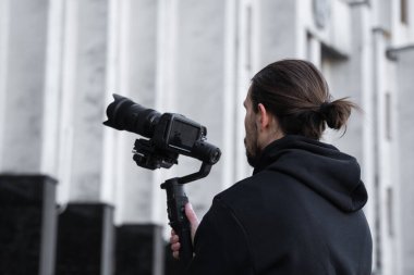 3 eksenli titreşimli dengeleyicide profesyonel kamera tutan genç profesyonel kameraman. Pro ekipmanlar titremeden yüksek kaliteli video çekmeye yardımcı olur. Siyah kapüşonlu bir kameraman video çekiyor..