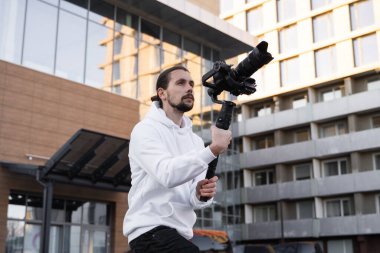 3 eksenli titreşimli dengeleyicide profesyonel kamera tutan genç profesyonel kameraman. Pro ekipmanlar titremeden yüksek kaliteli video çekmeye yardımcı olur. Beyaz kapüşonlu bir kameraman video çekiyor..