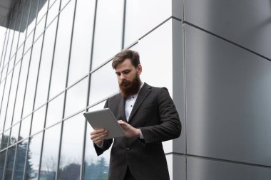 Modern genç sakallı iş adamı dijital tabletle çalışıyor. Genç hippi işadamı tableti açık havada tutuyor. Bir ofis binasının önünde dururken bir tabletle çevrimiçi çalışmak.