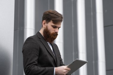 Modern genç sakallı iş adamı dijital tabletle çalışıyor. Genç hippi işadamı tableti açık havada tutuyor. Bir ofis binasının önünde dururken bir tabletle çevrimiçi çalışmak.