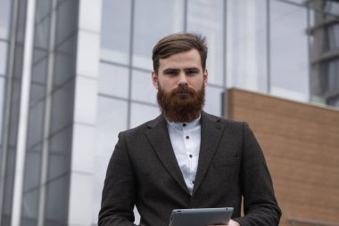 Modern genç sakallı iş adamı elinde dijital tabletle ayakta duruyor. Genç hippi işadamı tableti açık havada tutuyor. Bir ofis binasının önünde dururken bir tabletle çevrimiçi çalışmak.