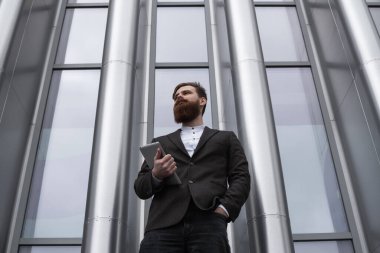 Modern genç sakallı iş adamı elinde dijital tabletle ayakta duruyor. Genç hippi işadamı tableti açık havada tutuyor. Bir ofis binasının önünde dururken bir tabletle çevrimiçi çalışmak.