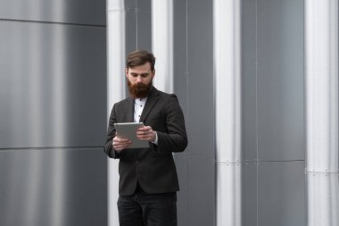 Modern genç sakallı iş adamı dijital tabletle çalışıyor. Genç hippi işadamı tableti açık havada tutuyor. Bir ofis binasının önünde dururken bir tabletle çevrimiçi çalışmak.