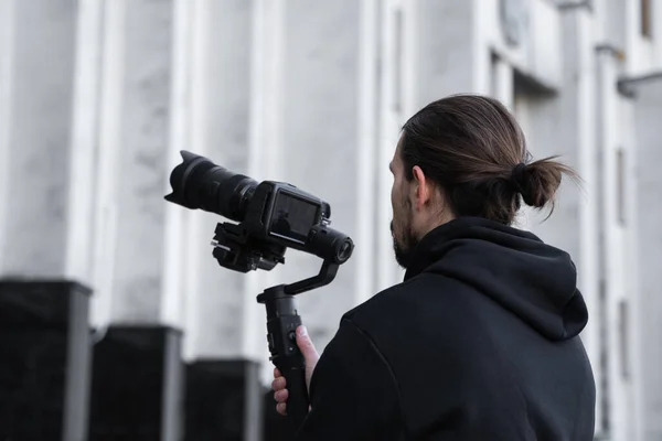 Young Professional videographer holding professional camera on 3-axis gimbal stabilizer. Pro equipment helps to make high quality video without shaking. Cameraman wearing black hoodie making a videos. — Stock Photo, Image