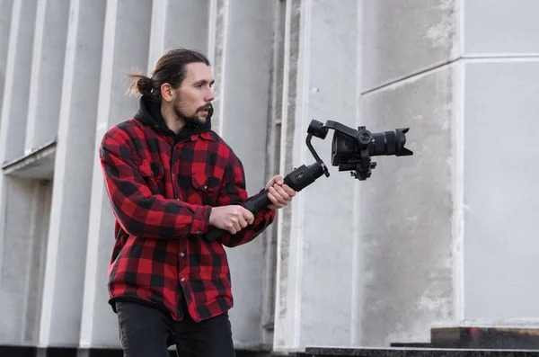 Νέοι επαγγελματίες videographer κρατώντας επαγγελματική φωτογραφική μηχανή σε 3-άξονα gimbal σταθεροποιητή. Pro εξοπλισμός βοηθά να κάνει υψηλής ποιότητας βίντεο χωρίς ανακίνηση. Καμεραμάν φορώντας κόκκινο πουκάμισο κάνοντας βίντεο. — Φωτογραφία Αρχείου