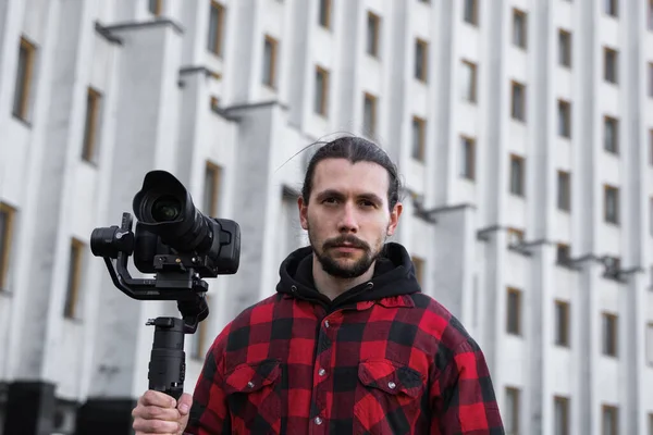 Jovem videógrafo profissional segurando câmera profissional no estabilizador de 3 eixos gimbal. Pro equipamento ajuda a fazer vídeo de alta qualidade sem tremer. Cameraman vestindo camisa vermelha fazendo um vídeos . — Fotografia de Stock