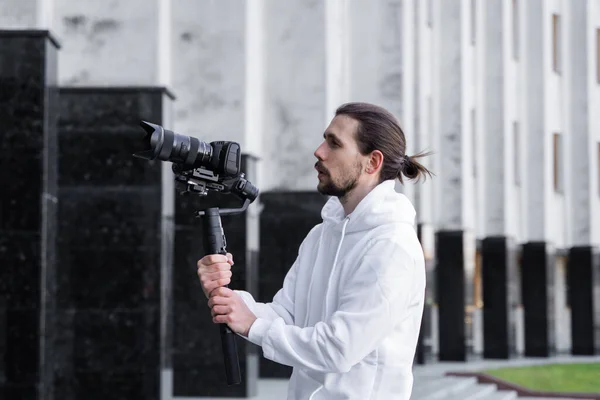 Giovane videografo professionale con fotocamera professionale su stabilizzatore gimbale a 3 assi. L'attrezzatura Pro aiuta a fare video di alta qualità senza scuotere. Cameraman indossa felpa bianca facendo un video . — Foto Stock