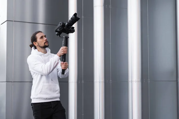 Fiatal profi videós professzionális kamerát tart 3 tengelyes gimbal stabilizátoron. Pro berendezések segít, hogy kiváló minőségű videót rázás nélkül. Az operatőr fehér kapucnis pulóvert visel, videókat készít. — Stock Fotó