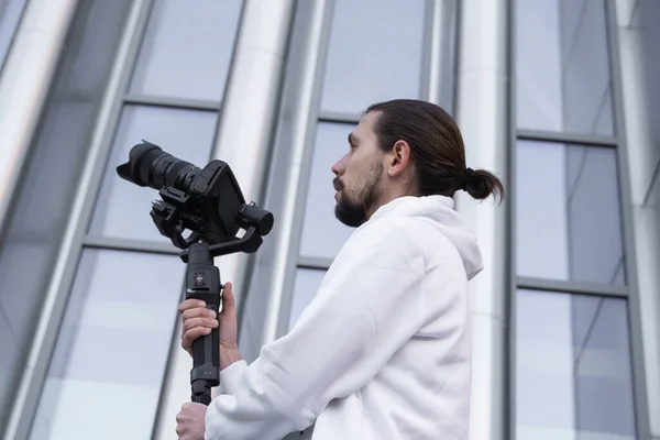 Νέοι επαγγελματίες videographer κρατώντας επαγγελματική φωτογραφική μηχανή σε 3-άξονα gimbal σταθεροποιητή. Pro εξοπλισμός βοηθά να κάνει υψηλής ποιότητας βίντεο χωρίς ανακίνηση. Καμεραμάν με άσπρη κουκούλα που φτιάχνει βίντεο. — Φωτογραφία Αρχείου