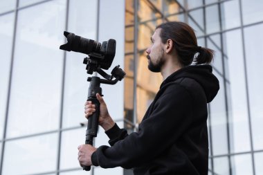 3 eksenli titreşimli dengeleyicide profesyonel kamera tutan genç profesyonel kameraman. Pro ekipmanlar titremeden yüksek kaliteli video çekmeye yardımcı olur. Beyaz kapüşonlu bir kameraman video çekiyor..