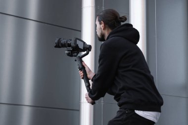 3 eksenli titreşimli dengeleyicide profesyonel kamera tutan genç profesyonel kameraman. Pro ekipmanlar titremeden yüksek kaliteli video çekmeye yardımcı olur. Beyaz kapüşonlu bir kameraman video çekiyor..