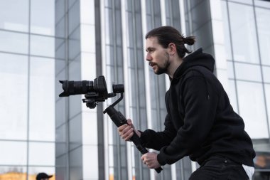 Young Professional videographer holding professional camera on 3-axis gimbal stabilizer. Pro equipment helps to make high quality video without shaking. Cameraman wearing white hoodie making a videos. clipart