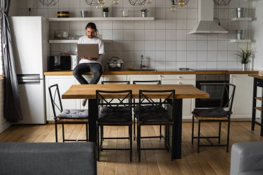 Evde çalışan serbest çalışan bir işçi mutfak atölyesinde oturuyor ve dizüstü bilgisayar kullanıyor. Laptopla çalışan ve haber okuyan sakallı bir adam. Yakışıklı, başarılı, modern evinde çalışan bir girişimci..