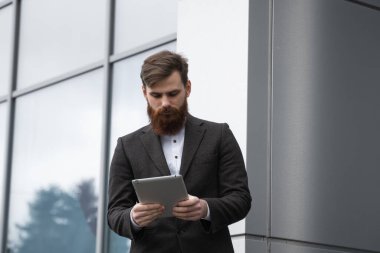 Modern genç sakallı iş adamı dijital tabletle çalışıyor. Genç hippi işadamı tableti açık havada tutuyor. Bir ofis binasının önünde dururken bir tabletle çevrimiçi çalışmak.