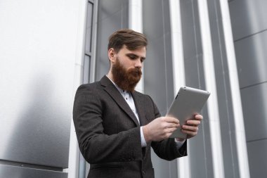 Modern genç sakallı iş adamı dijital tabletle çalışıyor. Genç hippi işadamı tableti açık havada tutuyor. Bir ofis binasının önünde dururken bir tabletle çevrimiçi çalışmak.