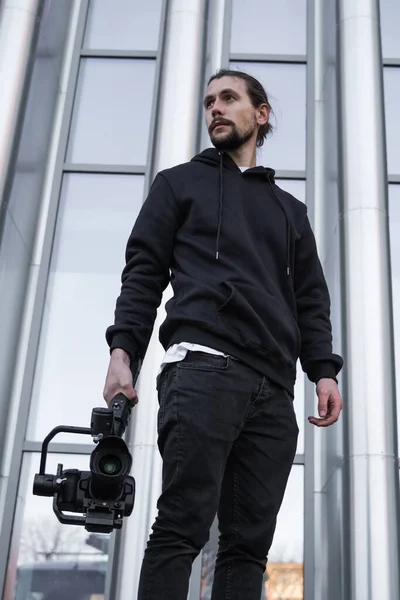 Jonge professionele videograaf met professionele camera op 3-assige gimbal stabilisator. Pro-apparatuur helpt om video van hoge kwaliteit te maken zonder te schudden. Cameraman draagt witte hoodie een video 's maken. — Stockfoto