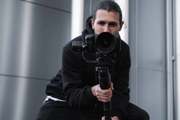 Fiatal profi videós professzionális kamerát tart 3 tengelyes gimbal stabilizátoron. Pro berendezések segít, hogy kiváló minőségű videót rázás nélkül. Az operatőr fehér kapucnis pulóvert visel, videókat készít. — Stock Fotó