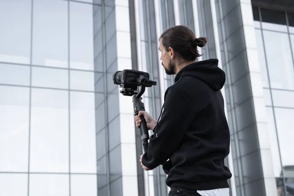 Young Professional videographer holding professional camera on 3-axis gimbal stabilizer. Pro equipment helps to make high quality video without shaking. Cameraman wearing white hoodie making a videos.