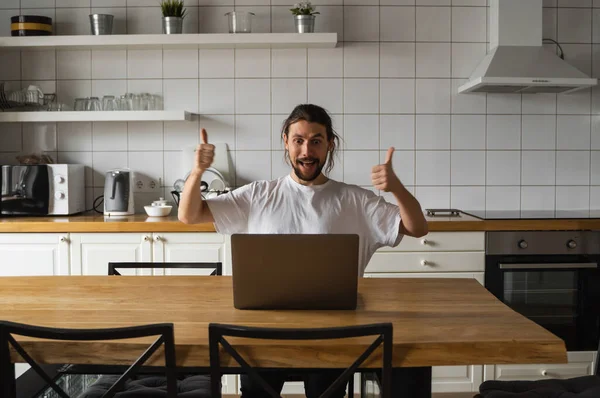 Ενθουσιασμένος επιχειρηματίας που χαίρεται για το επιτυχημένο έργο. Happy Man κρατώντας αντίχειρες στον αέρα και κοιτάζοντας φορητό υπολογιστή, ενώ κάθεται σε μια κουζίνα στο σπίτι. Ευτυχισμένος ελεύθερος επαγγελματίας, φοιτητής που εργάζεται σε φορητό υπολογιστή — Φωτογραφία Αρχείου