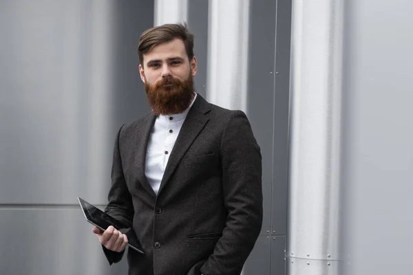 Modern Young barbudo hombre de negocios de pie con una tableta digital. Joven empresario hipster sosteniendo tableta en las manos al aire libre. Trabajar en línea con una tableta mientras está parado afuera en un edificio de oficinas . — Foto de Stock