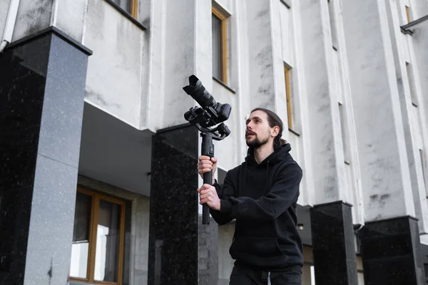 Young Professional videographer holding professional camera on 3-axis gimbal stabilizer. Pro equipment helps to make high quality video without shaking. Cameraman wearing black hoodie making a videos.