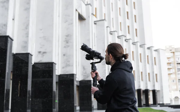 Young Professional videographer holding professional camera on 3-axis gimbal stabilizer. Pro equipment helps to make high quality video without shaking. Cameraman wearing black hoodie making a videos.