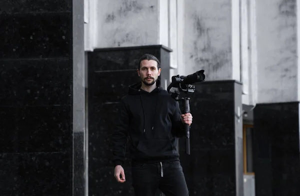 Νέοι επαγγελματίες videographer κρατώντας επαγγελματική φωτογραφική μηχανή σε 3-άξονα gimbal σταθεροποιητή. Pro εξοπλισμός βοηθά να κάνει υψηλής ποιότητας βίντεο χωρίς ανακίνηση. Καμεραμάν με μαύρη κουκούλα που φτιάχνει βίντεο. — Φωτογραφία Αρχείου