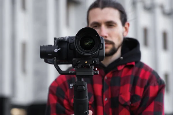 Giovane videografo professionale con fotocamera professionale su stabilizzatore gimbale a 3 assi. L'attrezzatura Pro aiuta a fare video di alta qualità senza scuotere. Cameraman indossa camicia rossa facendo un video . — Foto Stock