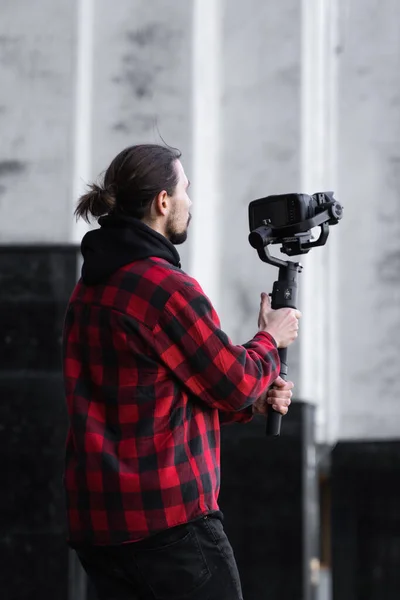 Jeune vidéaste professionnel tenant une caméra professionnelle sur stabilisateur de cardan 3 axes. L'équipement Pro aide à faire des vidéos de haute qualité sans trembler. Cameraman portant chemise rouge faisant une vidéo . — Photo