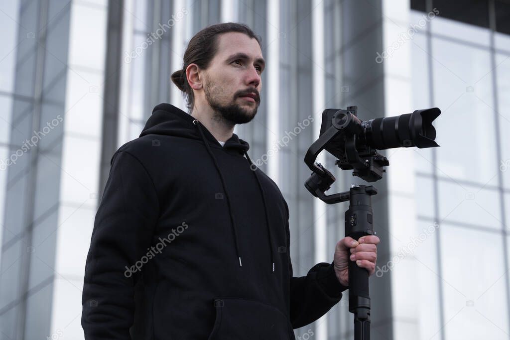 Young Professional videographer holding professional camera on 3-axis gimbal stabilizer. Pro equipment helps to make high quality video without shaking. Cameraman wearing white hoodie making a videos.