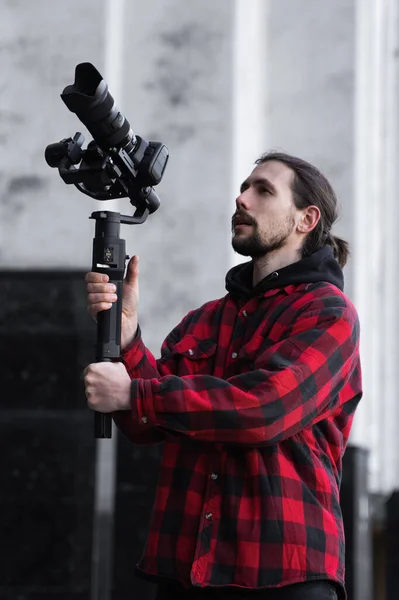 Jovem videógrafo profissional segurando câmera profissional no estabilizador de 3 eixos gimbal. Pro equipamento ajuda a fazer vídeo de alta qualidade sem tremer. Cameraman vestindo camisa vermelha fazendo um vídeos . — Fotografia de Stock