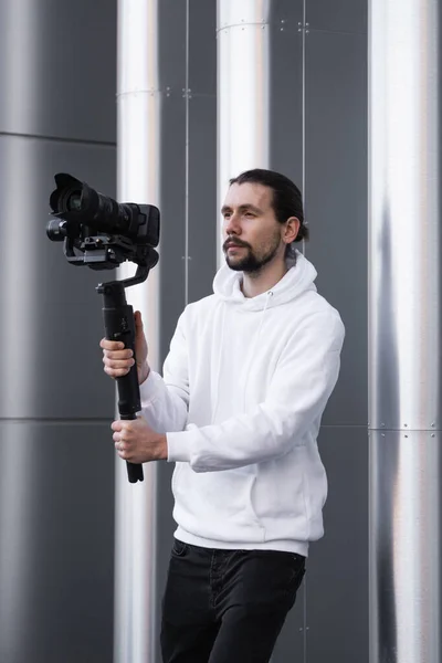 Ung professionell videograf som håller professionell kamera på 3-axlig gimbal stabilisator. Pro-utrustning hjälper till att göra högkvalitativ video utan att skaka. Kameraman bär vit luvtröja gör en video. — Stockfoto