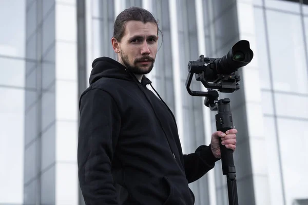 Young Professional videographer holding professional camera on 3-axis gimbal stabilizer. Pro equipment helps to make high quality video without shaking. Cameraman wearing white hoodie making a videos.