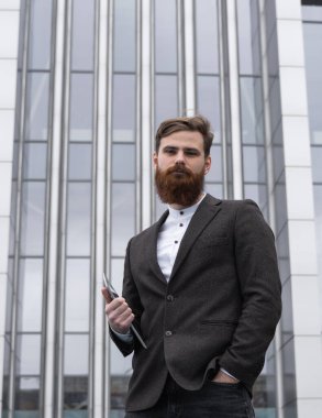 Modern genç sakallı iş adamı elinde dijital tabletle ayakta duruyor. Genç hippi işadamı tableti açık havada tutuyor. Bir ofis binasının önünde dururken bir tabletle çevrimiçi çalışmak.