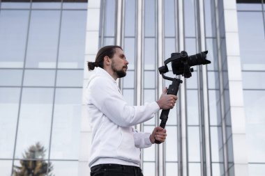 3 eksenli titreşimli dengeleyicide profesyonel kamera tutan genç profesyonel kameraman. Pro ekipmanlar titremeden yüksek kaliteli video çekmeye yardımcı olur. Beyaz kapüşonlu bir kameraman video çekiyor..