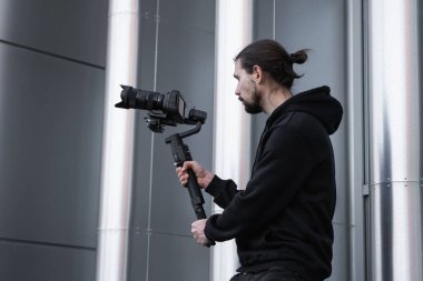 3 eksenli titreşimli dengeleyicide profesyonel kamera tutan genç profesyonel kameraman. Pro ekipmanlar titremeden yüksek kaliteli video çekmeye yardımcı olur. Beyaz kapüşonlu bir kameraman video çekiyor..