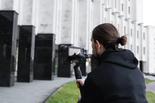 Jonge professionele videograaf met professionele camera op 3-assige gimbal stabilisator. Pro-apparatuur helpt om video van hoge kwaliteit te maken zonder te schudden. Cameraman dragen zwarte hoodie het maken van een video 's. — Stockfoto